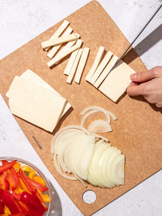 knife slicing tofu into matchsticks on wood cutting board with sliced onions and peppers