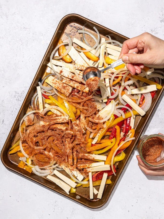 hand shaking seasoning over sliced tofu and peppers on baking sheet