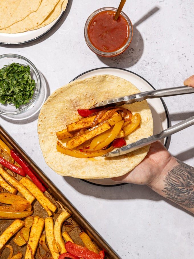 hand with placing tofu onto tortilla with tongs beside salsa and cilantro