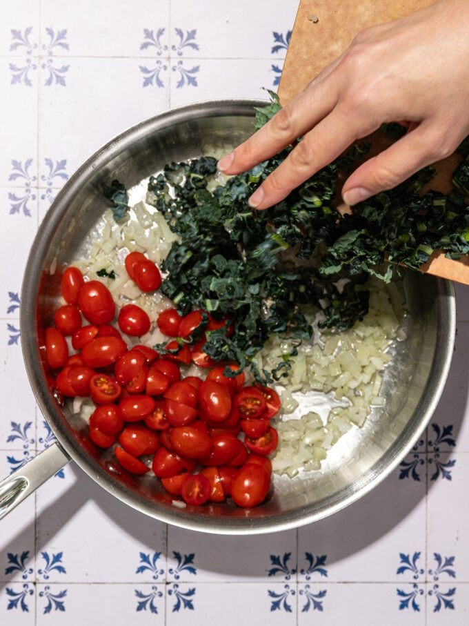 hand adding kale to skillet with cherry tomatoes and onion
