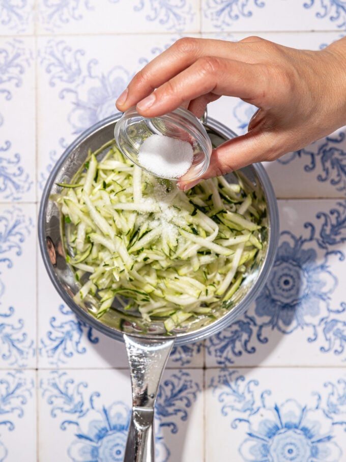 hand pouring salt onto shredded zucchini in a colander 