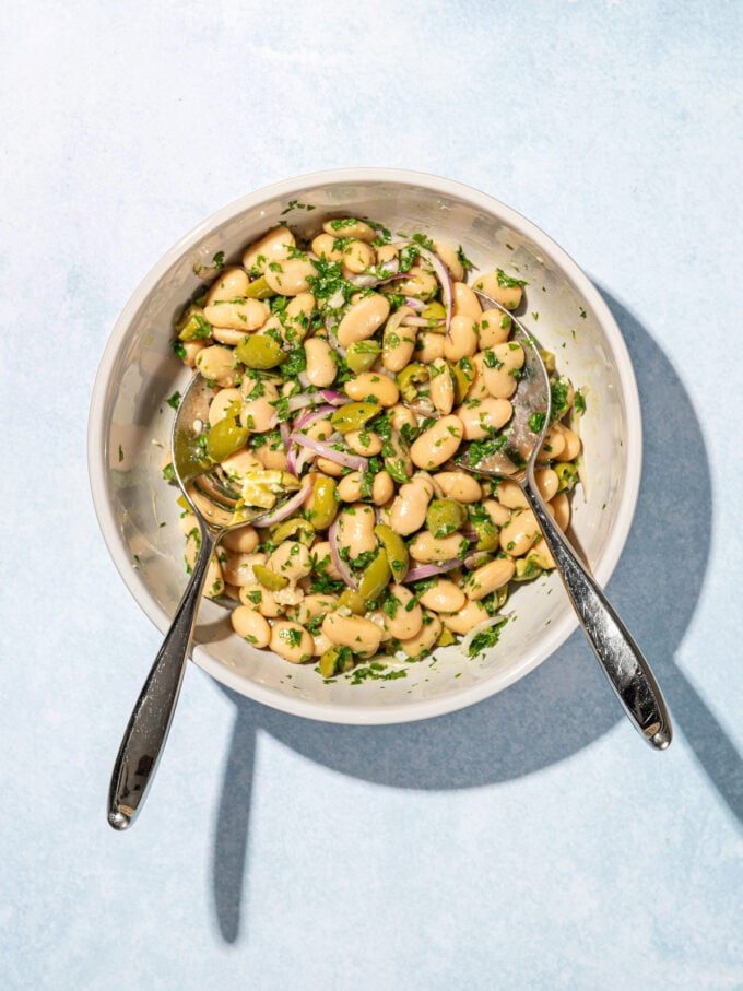 white bowl filled with marinated white bean, onion and olive salad, with two salad servers resting on top