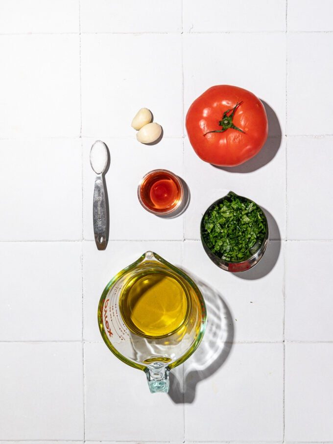 glass cup of olive oil beside cloves of garlic, a tomato, minced parsley, salt and vinegar