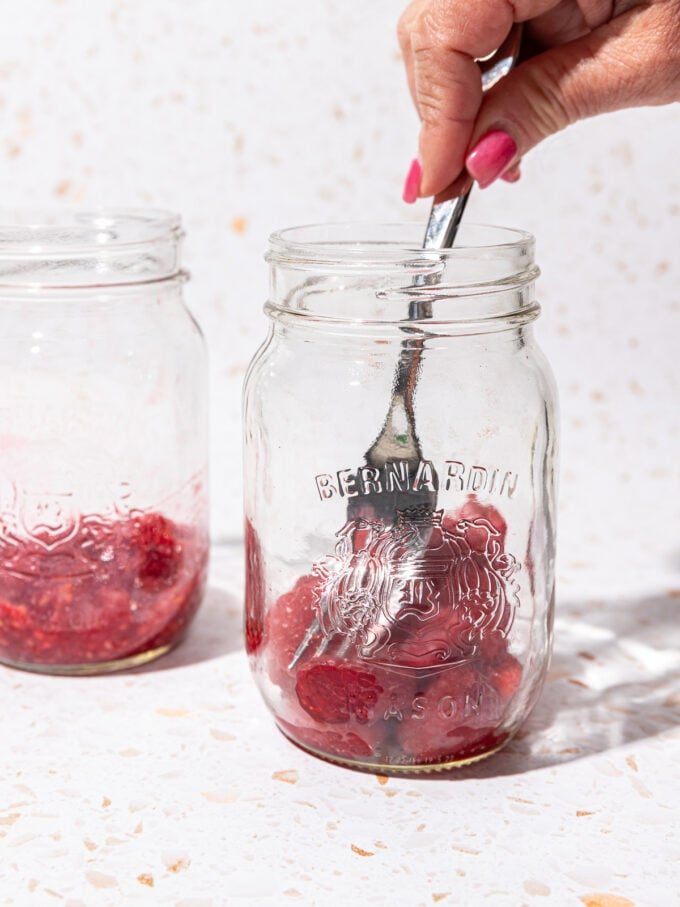 hand mashing raspberries in a mason jar with a fork