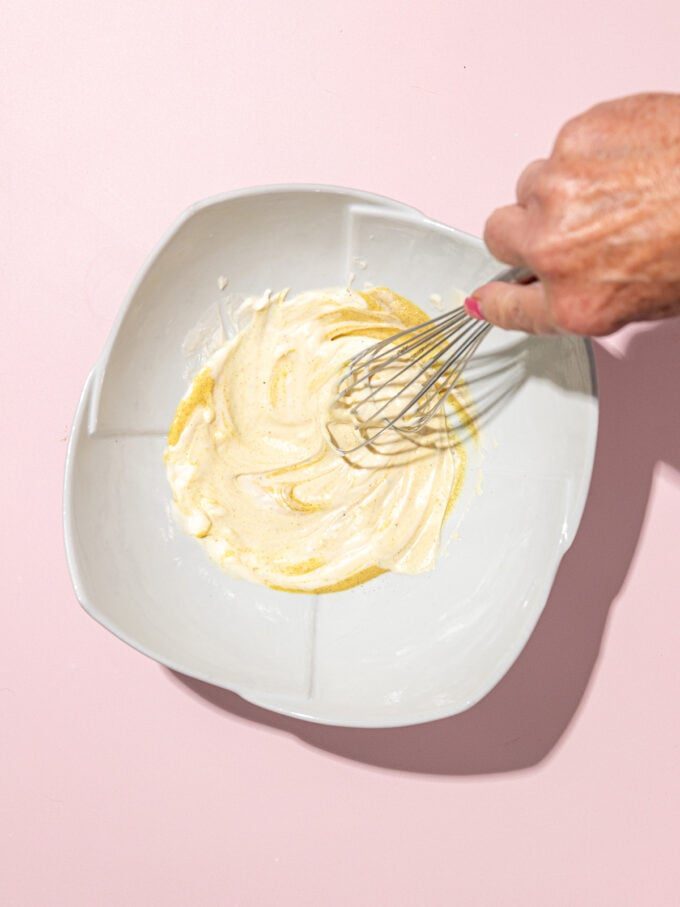 hand whisking mayo and mustard in white bowl on pink background