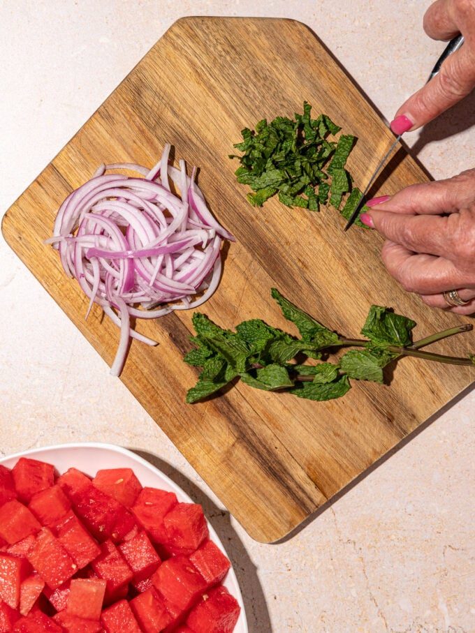 hand slicing mint leaves on wooden cutting board with sliced red onion beside it