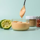 hand holding spoon of cashew dressing over glass jar of dressing, with spent limes, and jars of cashew butter and chile flakes beside it
