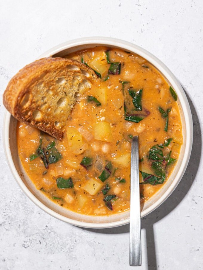 white bean stew in a white bowl with a spoon and slice of toast dunked in
