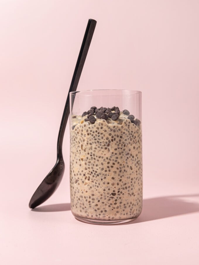 glass filled with cookie dough overnight oats with a black spoon leaned up against it, on a pink background