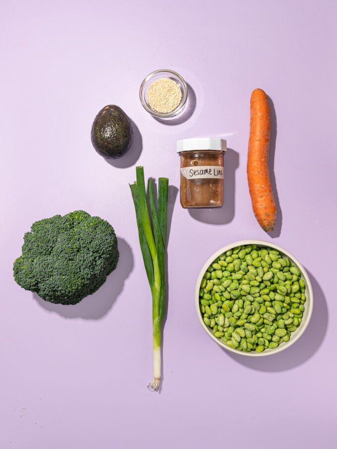 bowl of edamame, a carrot, broccoli, green onion, avocado and jar of dressing on purple background